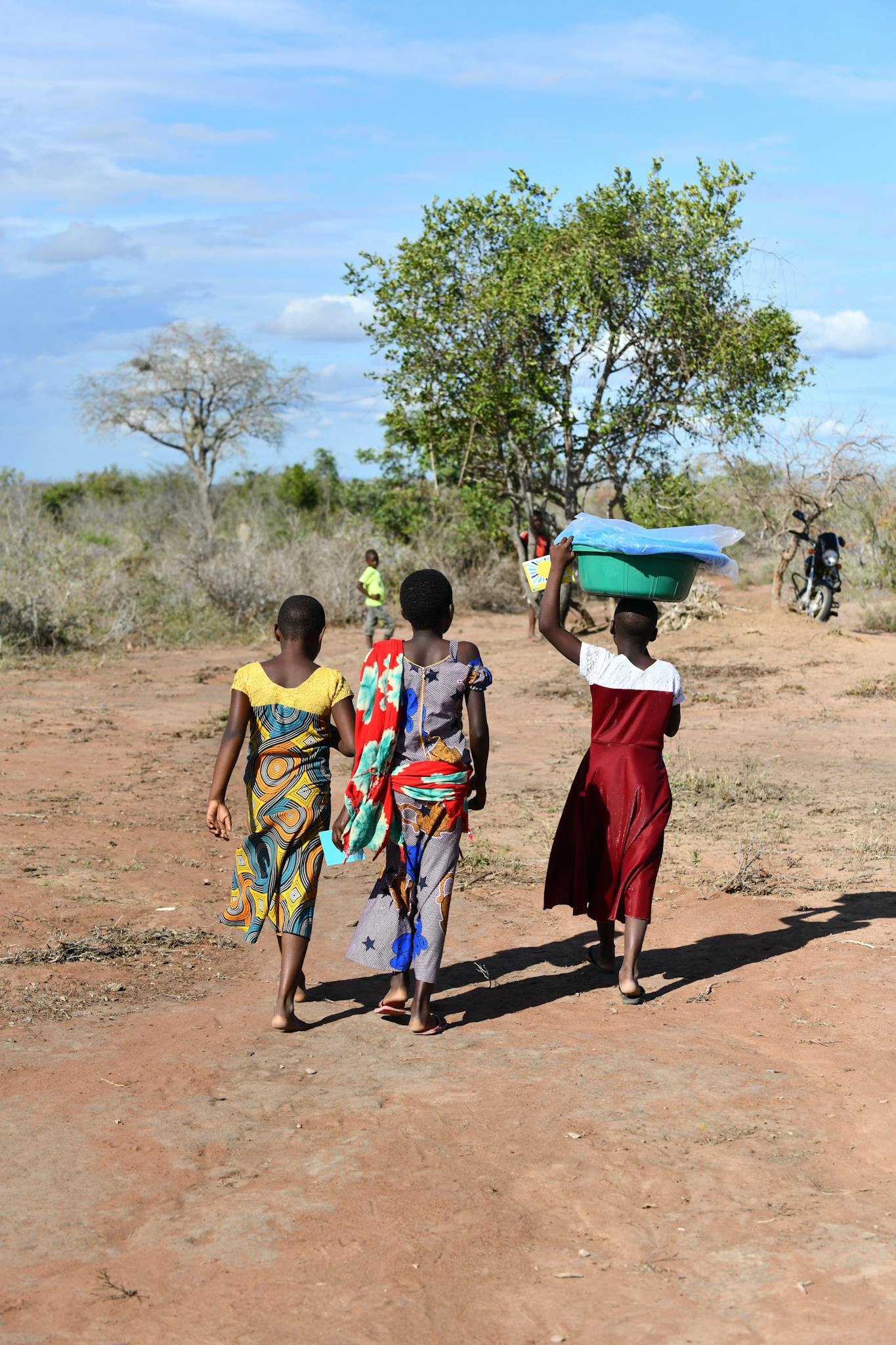 Three African girls walking on a rural path, carrying supplies on a sunny day.